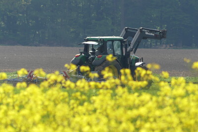 Rapsblüte mit Schlepper im Hintergrund - © Hendrik Hillebrand Ein Traktor pflügt ein Feld, im Vordergrund blühende gelbe Blumen, im Hintergrund ein Wald.
