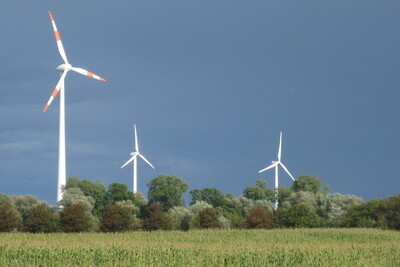 Der Austausch von 2 %-Punkten Braunkohlestrom durch Windstrom bringt in Deutschland so viel Treibhausgaseinsparung wie eine Halbierung des Fleischkonsums - © Ansgar Lasar Mehrere Windräder stehen auf einem Feld vor einem dunklen, bewölkten Himmel.