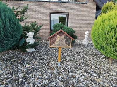 So lieber nicht - Insektenhotel in einem Schottergarten - © Nadja Krause Ein Insektenhotel steht in einem mit Kies bedeckten Garten, umgeben von Büschen und kleinen Statuen, vor einem Backsteingebäude.